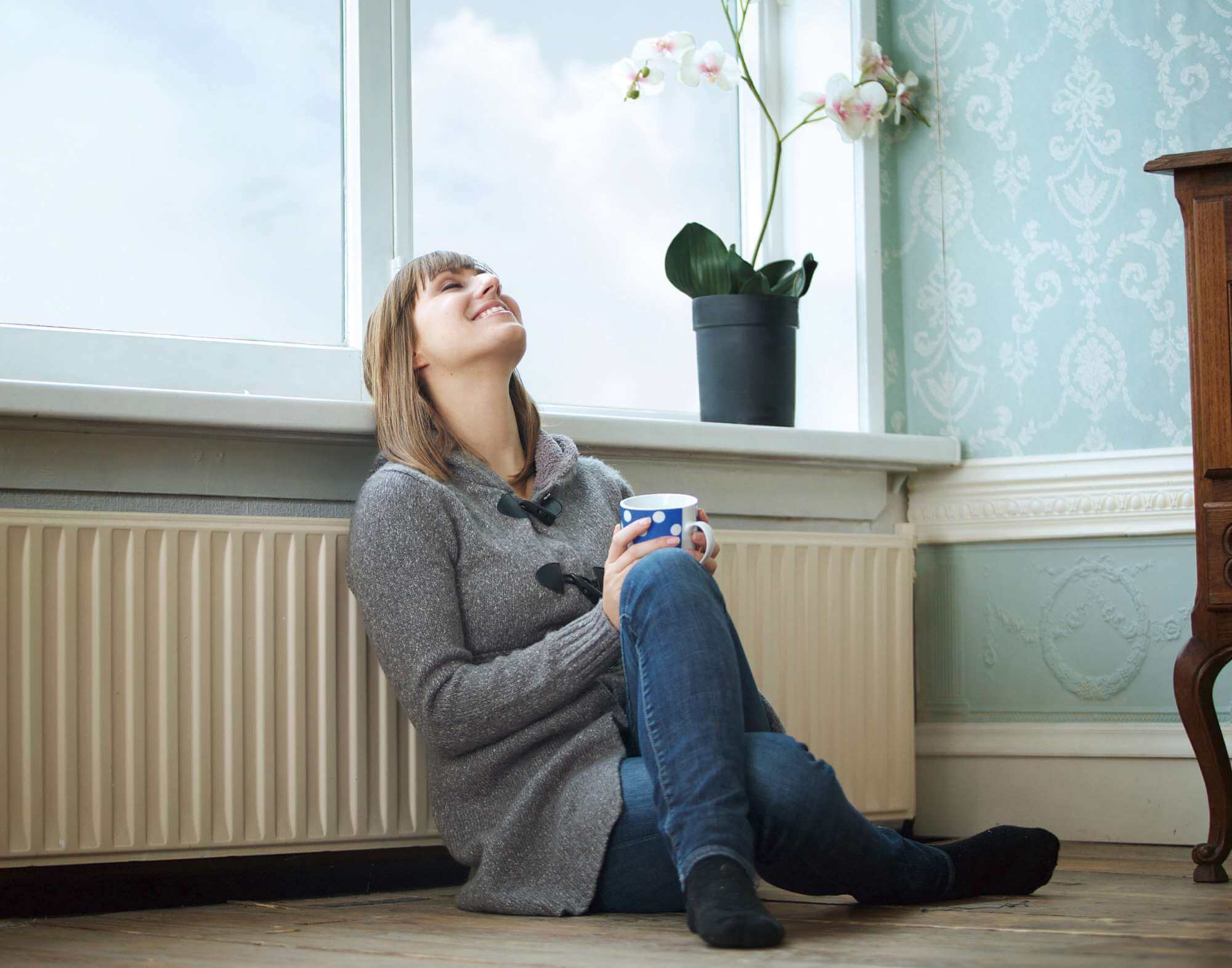 Kvinde sidder op ad radiator med en kop i hånden